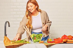 Woman preparing fresh salad in kitchen beside Kencko Smoothie ingredients.